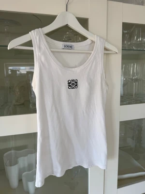 Vit ribbad linnetopp från Loewe - Säljer en vit ribbad linnetopp från Loewe med svart broderad logga på bröstet. Toppen är ärmlös och har en klassisk rund halsringning. Perfekt att styla till jeans eller kjol för en clean och trendig look. Anagram tank top in cotton. En liten fläck som knappt syns, se bild 4, fint använt skick. Köpt second hand.