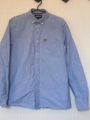 Ljusblå skjorta från Lyle & Scott - Klassisk ljusblå skjorta från Lyle & Scott med knappslå och bröstficka. Skjortan har lång ärm, button down-krage och den ikoniska gula loggan broderad på bröstet. Tillverkad i mjuk bomull för en bekväm känsla.