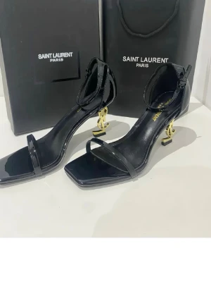 Svarta YSL pumps med gulddetalj - Säljer ett par svarta pumps från Saint Laurent med öppen tå och justerbar ankelrem. Klackarna är i guld med ikoniska YSL-loggan som design. Skorna är i lackat läder och har en elegant, modern siluett som verkligen sticker ut. Låda och kasse tillkommer ej.