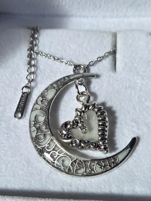 Halsband med mån- och hjärthänge - självlysande  - Unikt handgjort halsband i silverfärgad metall med ett halvmåneformat hänge dekorerat med vita detaljer och ett hjärta i mitten. Hänget lyser i mörkret med ett starkt blått sken efter att ha varit i vanligt dagsljus utifrån eller solljus en kort stund. Kedjan är tunn och justerbar i rostfritt stål. Perfekt för dig som gillar magiska och fantasifulla smycken. Både hänget och kedjan tål vatten utan att missfärgas. Hängets storlek ca 3 cm. Kedjan 45 cm + 5 cm förlängningskedja. 