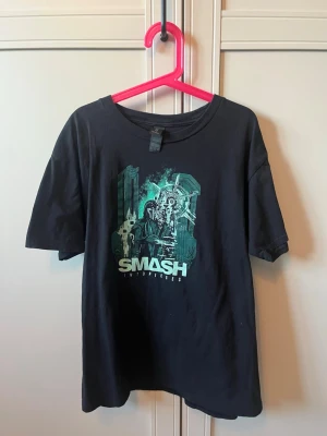Svart Smash T-shirt från Gildan - Cool svart T-shirt från smash into pieces med ett futuristiskt grönt och blått tryck på bröstet på apoc och texten 'SMASH INTO PIECES'. Tillverkad i mjuk bomull och har en klassisk passform med korta ärmar. Perfekt för dig som gillar unika grafiska motiv.