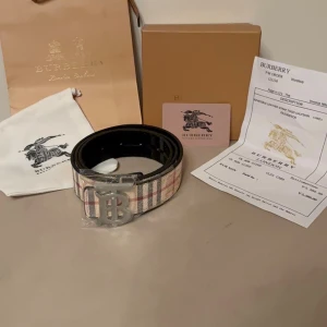 Randigt Burberry bälte med metallspänne - Säljer ett klassiskt bälte med det ikoniska randiga mönstret i beige, svart, rött och vitt. Bältet har ett stort metallspänne med logotyp och insida i svart skinn. Perfekt accessoar för att lyfta vilken outfit som helst.