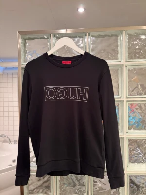Svart sweatshirt från Hugo Boss - Snygg svart sweatshirt från Hugo Boss med vit HUGO-logga tryckt i spegelvänd stil på bröstet. Tröjan har rund halsringning, långa ärmar och ribbade muddar vid ärmslut och nederkant. Perfekt för en stilren och avslappnad look.