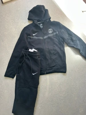 Nike PSG Strike Tech Fleece Tracksuit - Stilrent Nike PSG Strike Tech Fleece tracksuit i svart 🖤 Set med överdel och byxor i bra skick och fin kvalitet. Mjukt material och tydliga PSG-detaljer ger en clean och sportig helhet. Passar storlek S längd (170-176)Snabb frakt 📦