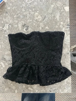 Svart spets korsett peplum - Snygg svart korsettopp med peplum och spetsdetaljer. Axelbandslös design och markerade kupor ger en feminin siluett. Perfekt att styla med höga jeans eller kjol för en trendig look.