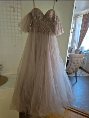 Ljusbeige långklänning från Queen - Magisk långklänning i ljusbeige från Queen Brud & Fest. Klänningen har glittrig överdel med pärldetaljer, v-ringning och puffiga korta ärmar. Kjolen är i flera lager tunn tyll som ger en drömmig look. Perfekt för bal eller festliga tillfällen. Dragkedja är trasig på den måste bytas och snöret har jag tappat 