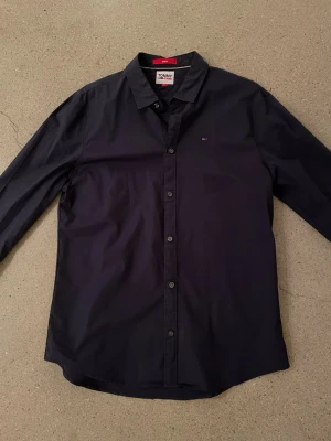 Mörkblå skjorta från Tommy Jeans - Snygg mörkblå skjorta från Tommy Jeans i slim fit-modell. Skjortan har klassisk krage, knappar framtill och en liten broderad logga på bröstet. Tillverkad i mjuk bomull som känns skön mot huden. Perfekt för en clean och stilren look.