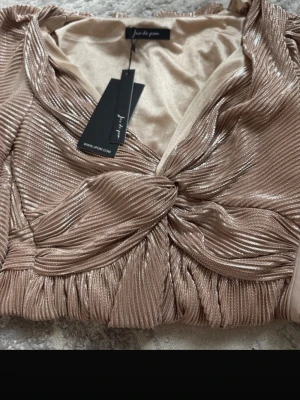 Glittrig champagnefärgad klänning  Jus de Pom - Supersnygg glittrig klänning från Jus de Pom i champagnefärg med twistad detalj framtill. Plisserad struktur och v-ringad halsringning ger en lyxig känsla. Perfekt för dig som vill sticka ut lite extra.