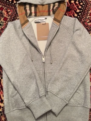 Grå zip hoodie från Burberry - Snygg grå zip hoodie från Burberry med klassiskt rutigt foder i huvan och dragsko. Tillverkad i mjuk bomullsblandning, har fickor på sidorna och diskret Burberry-logga broderad på bröstet. Perfekt för en chill och stilren look.
