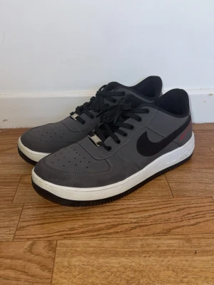 Nike Air Force 1 grå sneakers - Snygga Nike Air Force 1 sneakers i grått med svart swoosh och vita sulor. De har använda några gånger men inte förstörda någon stans. Skorna har svarta skosnören, perforerad tåbox och diskreta röda detaljer på sidan. Ovandelen är i syntetmaterial och mesh för extra komfort. Perfekta för en clean och stilren look.