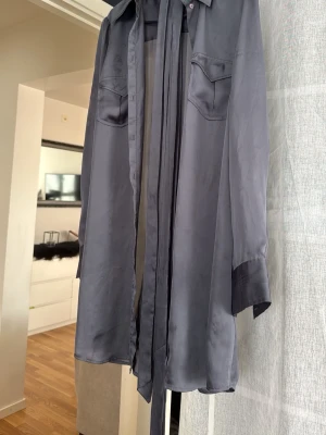 Mörkgrå sidenblus med knytband - Snygg mörkgrå blus i glansigt sidenmaterial med långa ärmar, två bröstfickor och ett långt knytband vid kragen. Man kan även knyta runt midjan och ha det som en kort klänning. Passar även M/S