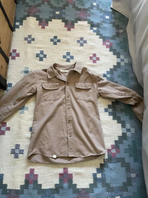 Beige skjorta från I dig denim - Säljer en beige skjorta från I dig denim med klassisk krage, två bröstfickor och knappar framtill. Skjortan har lång ärm och är tillverkad i mjuk bomull, perfekt för en avslappnad och stilren look. Den har ett litet hål och slitningar på baksidan från skating