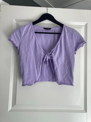 Ljuslila croppad topp med knyt - Säljer en ljuslila croppad topp med korta ärmar och v-ringning. Toppen har en snygg knytning framtill som ger en trendig look. Materialet känns mjukt och är troligtvis bomull eller en bomullsblandning. Perfekt att styla med jeans eller kjol för en chill vibe.