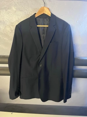 Marinblå kavaj med 2 knappars enkelknäppning - Stilren marinblå herrkavaj med enkelknäppning och två framfickor. Kavajen har klassiska slag och är helfodrad för en snygg siluett. Perfekt till skjorta eller t-shirt för en clean look. Materialet känns slätt och har en lätt glans.