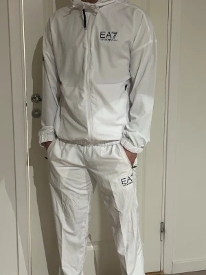 Vit EA7 träningsdress med huva - Säljer en stilren vit träningsdress från EA7 Emporio Armani. Setet består av en jacka med dragkedja, huva och svarta detaljer samt matchande byxor med resår i midjan. Båda plaggen har EA7-logga tryckt på bröstet och benet. Perfekt för sport eller chill. Den har endast en liten defekt vid sidan av byxorna. 