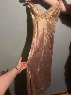 Champagnefärgad långklänning med spets - Säljer en champagnefärgad långklänning med tunna axelband och spetsdetaljer vid bysten. Klänningen har omlottknytning i ryggen och är gjord i ett glansigt material.🫶🏻