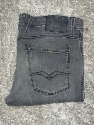 Replay Anbass jeans  - Säljer ett par gråa slim jeans från Replay med klassisk femficksdesign och diskret logga på bakfickan. Jeansen har smal passform och är tillverkade i stretchigt material för extra komfort. Perfekta för en trendig och avslappnad look. JAG SKICKAR EJ FLER BILDER PÅ JEANSEN! för storleksguide hänvisar jag till måtten och för bättre inblick i passformen rekommenderar jag att googla på modellnamnet. :) 1.102