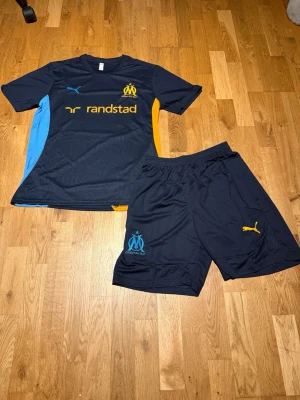 Marseille summer set navy blue/orange - Säljer ett Marseille fotbollskit från Puma med matchtröja och shorts. Setet är mörkblått med gula och ljusblå detaljer, Marseille-logga och Puma-logga på både tröja och shorts. Materialet är lätt och andas, perfekt för träning eller match.