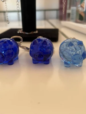 Blå nyckelringar grisar - Tre nyckelringar formade som små grisar i genomskinlig blå och ljusblått. Två av grisarna är mörkblå och en är ljusblå, alla med metallring för att fästa på nycklar eller väska. Texten 'NORDEA' syns på ovansidan av grisarna.