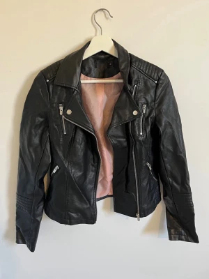 Svart biker skinnjacka med dragkedjor - Snygg svart skinnjacka i bikerstil med flera silvriga dragkedjor och detaljer och rosa innerfoder. Endast använd ca 4 gånger 