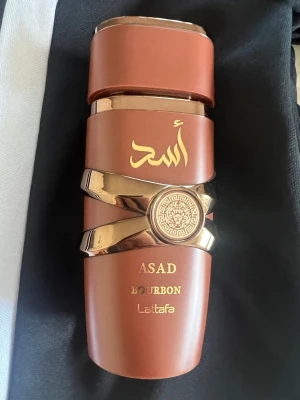 Lattafa Asad Bourbon parfym - Lattafa Asad Bourbon är en parfym i en brun, cylinderformad flaska med gulddetaljer och arabiska inskriptioner. Flaskan har en unik design med guldfärgade band och ett dekorativt emblem i mitten. Passar dig som gillar exklusiva och stilrena dofter. 80-90ml kvar av den