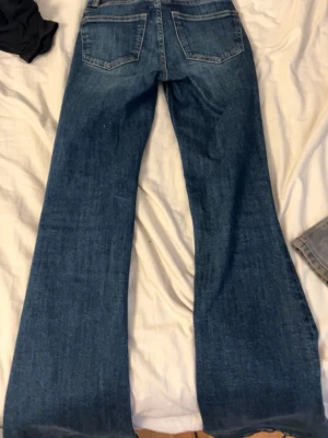 Blå bootcut jeans med medelhög midja - Säljer ett par klassiska blå bootcut jeans med medelhög midja och utsvängda ben. Jeansen har fem fickor och är gjorda i ett slitstarkt denimtyg. Perfekta för dig som gillar retrovibbar och vill ha en snygg siluett.