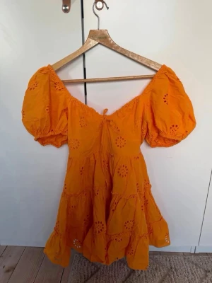 Orange kort klänning med puffärm - Säljer en superfin orange kort klänning med puffärmar och broderade blommönster. Klänningen har omlottkänsla upptill, volanger i kjolen och en söt knytning vid bysten. Perfekt för sommarens alla tillfällen!