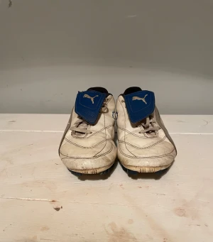 Puma Esito vita fotbollsskor - Säljer ett par vita Puma Esito fotbollsskor med blå detaljer och klassisk design. Skorna har blått snörskydd med Puma-logga, svarta innersidor och dobbar för spel på gräs. Tillverkade i slitstarkt syntetmaterial..