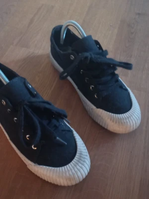 Svarta låga tygsneakers - Klassiska svarta sneakers i tyg med vita gummisulor och tåparti. Skorna har svarta skosnören och silverfärgade öljetter. Perfekta för en avslappnad och stilren look.