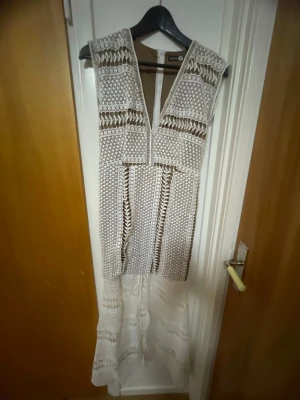 Vit långklänning med spetsdetaljer - Säljer en vit långklänning med v-ringning och vackra spetsdetaljer. Klänningen är ärmlös och har ett bohemiskt mönster med inslag av genombruten spets. Perfekt för dig som vill ha en unik och elegant look.