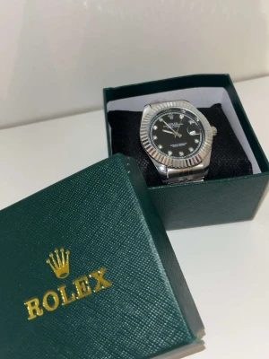 Rolex Datejust silver klocka - Snygg Rolex Datejust klocka i silver med svart urtavla och jubilee-länk. Klockan har markerade index med stenar och räfflad bezel. Kommer i original grön Rolex-box. Perfekt accessoar för dig som gillar klassisk och lyxig stil.