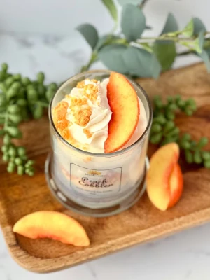 Sojaljus Peach Cobbler i glas - Unikt doftljus i glas med design som liknar en persikodessert. Ljuset är vitt med lager av persikofärgade detaljer och dekorerat med en konstgjord persikoskiva och gräddliknande topp. Tillverkat av 100% naturlig sojavax och har etikett med texten 'Peach Cobbler'.