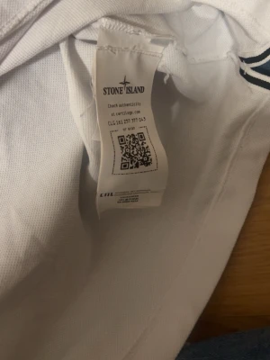 Vit pikétröja från Stone Island - Snygg vit pikétröja från Stone Island med klassisk krage och två svarta knappar. Tröjan har korta ärmar och en broderad logotyp på bröstet. Tillverkad i mjuk bomull som känns skön mot huden. Perfekt för en clean och stilren look.