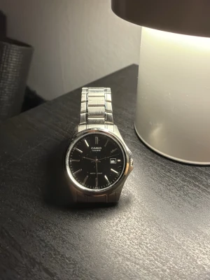 Casio klocka med svart urtavla - Stilren Casio armbandsklocka med svart urtavla och datumvisning. Klockan har ett klassiskt runt boett i silverfärgad metall och matchande länkarmband i rostfritt stål. Perfekt accessoar för dig som gillar enkel och tidlös design.