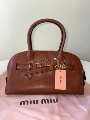 Brun handväska från Miu Miu - Snygg brun handväska från Miu Miu i slätt läder med guldfärgade metalldetaljer och dubbla handtag. Väskan har en rundad form, dragkedja upptill och diskret logotyp på framsidan. Perfekt accessoar för dig som gillar stilrena och exklusiva detaljer.