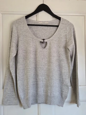 Ljusgrå stickad tröja Uterque - Snygg ljusgrå stickad tröja från Uterque med rund halsringning och unik cut-out detalj framtill som hålls ihop av en dekorativ ring. Tröjan har långa ärmar och ribbade muddar vid ärmslut och nederkant. Perfekt för en stilren och trendig look.