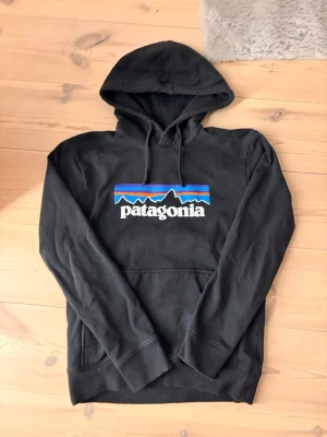 Svart Patagonia hoodie - Säljer en svart hoodie från Patagonia med det klassiska färgglada logotrycket på bröstet. Hoodien har huva med dragsko och en stor magficka. Tillverkad i mjukt bomullsmaterial som är skönt att ha på sig. Perfekt för dig som gillar streetwear och outdoor-vibe.