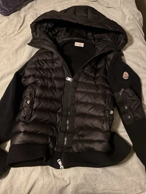 Svart cardigan  med huva från Moncler - Snygg svart dunjacka från Moncler med quiltad kropp, ribbade ärmar och muddar. Jackan har en stor huva, dragkedja framtill, fickor med knappar och en Moncler-logga på ärmen. Perfekt för kyliga dagar och riktigt trendig stil.