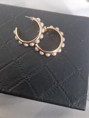 Guldfärgade hoops med pärlor - Säljer ett par snygga guldfärgade hoop-örhängen dekorerade med vita pärlor runt hela ringen. Örhängena har en klassisk rund form och ger en trendig touch till din stil. Perfekta för dig som vill ha något extra till din outfit.