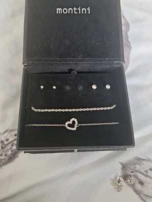 Silverfärgat armband med hjärta Montini - Snyggt silverfärgat armband från Montini med en kedja och ett hjärta dekorerat med små pärlor. Perfekt accessoar för att lyfta din outfit med en söt och trendig detalj. Kommer i original ask. Det kommer med två örhängen men den tredje är tyvärr borta. Dessa är aldrig använda förutom hjärtliga armbandet som är använd endast en gång