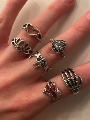 Set med 6 silvriga ringar med motiv - Säljer ett set med 6 unika silvriga ringar i olika former och motiv, bland annat hjärta, lotusblomma, skelett-hand, hjort och orm med röda detaljer. Ringarna är i metall och har en cool, bohemisk och alternativ stil. Helt oanvända och ej äkta silver, 7kr styck och alla för 40kr 🫶