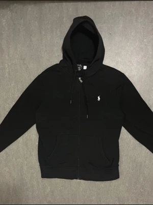 Äkta Polo Ralph Lauren Zip-up - Klassisk svart hoodie från Polo Ralph Lauren med dragkedja framtill, två fickor och justerbar huva. Ikoniska vita Polo-loggan broderad på bröstet. Tillverkad i mjukt bomullsmaterial som är skönt att bära. Perfekt för en avslappnad och stilren look.