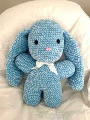 Stor blå virkad kanin med rosett - Virkat gosedjur i chenillegarn. Kaninen är 40cm lång och tillverkad för hand , vilket gör varje kanin unik.  Kaninen har svarta säkerhetsögon som sitter fast från insidan. Rekommenderas dock inte för de allra minsta barnen, då det alltid finns en viss risk, vid hårt drag, att ögonen lossnar.  Vitt satinband runt halsen som enkelt kan tas av.  Perfekt som present eller mysig inredningsdetalj!  Tvättråd: max 30°C, handtvätt.