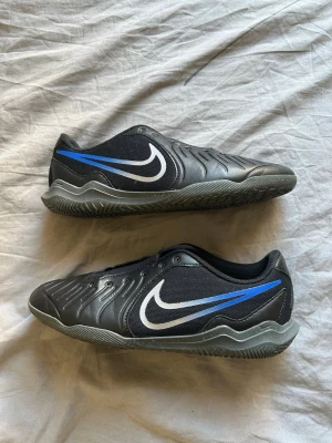 Nike svart/blå fotbollsskor - Säljer ett par svarta fotbollsskor från Nike med blå och vita detaljer. Skorna har en låg profil och är tillverkade i syntetmaterial med en vågig struktur på ovansidan. Den klassiska Nike-loggan syns tydligt på sidan och hälen har en unik symbol.