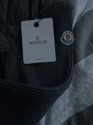 Grå cardigan från Moncler - Snygg grå dunväst från Moncler med ribbstickade detaljer på krage och ärmhål. Västen har dragkedja framtill, två sidofickor och Moncler-logga på axeln. Perfekt för lager-på-lager och ger en clean, modern look.
