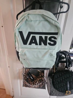 Mintgrön ryggsäck från Vans - Snygg mintgrön ryggsäck från Vans med stort svart logotryck på framsidan. Klassisk rundad form med en stor ytterficka och justerbara axelband. Tillverkad i slitstarkt tygmaterial, perfekt för skolan eller stan.