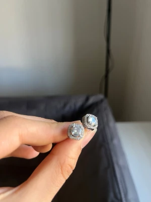 Silvriga örhängen med pärlor - Säljer ett par eleganta silvriga örhängen med runda vita pärlor i mitten, omgivna av gnistrande små stenar i en fyrkantig infattning. Perfekta för dig som gillar klassisk och stilren design. Passar till många olika looks och ger en lyxig touch.