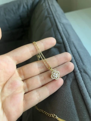 Guldhalsband med blomformad pärlberlock - Snyggt halsband i guld med en liten blomformad berlock dekorerad med vita pärlor. Kedjan är tunn och elegant, perfekt för att ge en söt touch till din outfit. Berlocken har en glittrig look tack vare pärldetaljerna.