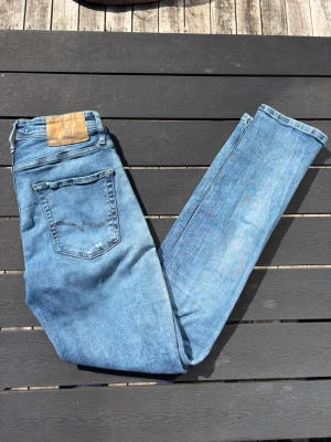 Blå jeans från Jack & Jones - Säljer ett par klassiska blå jeans från Jack & Jones med femficksmodell och ljus tvätt. Jeansen har raka ben och detaljerade sömmar på bakfickan. Tillverkade i bomull med en snygg läderpatch bak i midjan. Perfekta för en avslappnad och trendig stil.