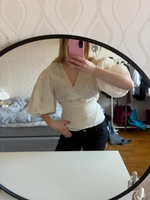 Vit omlottblus med puffärm - Säljer en vit omlottblus med djup v-ringning och stora puffärmar. Blusen har en feminin passform och är tillverkad i ett lätt, strukturerat tyg som ger en romantisk känsla. Perfekt att styla med både jeans och kjol.